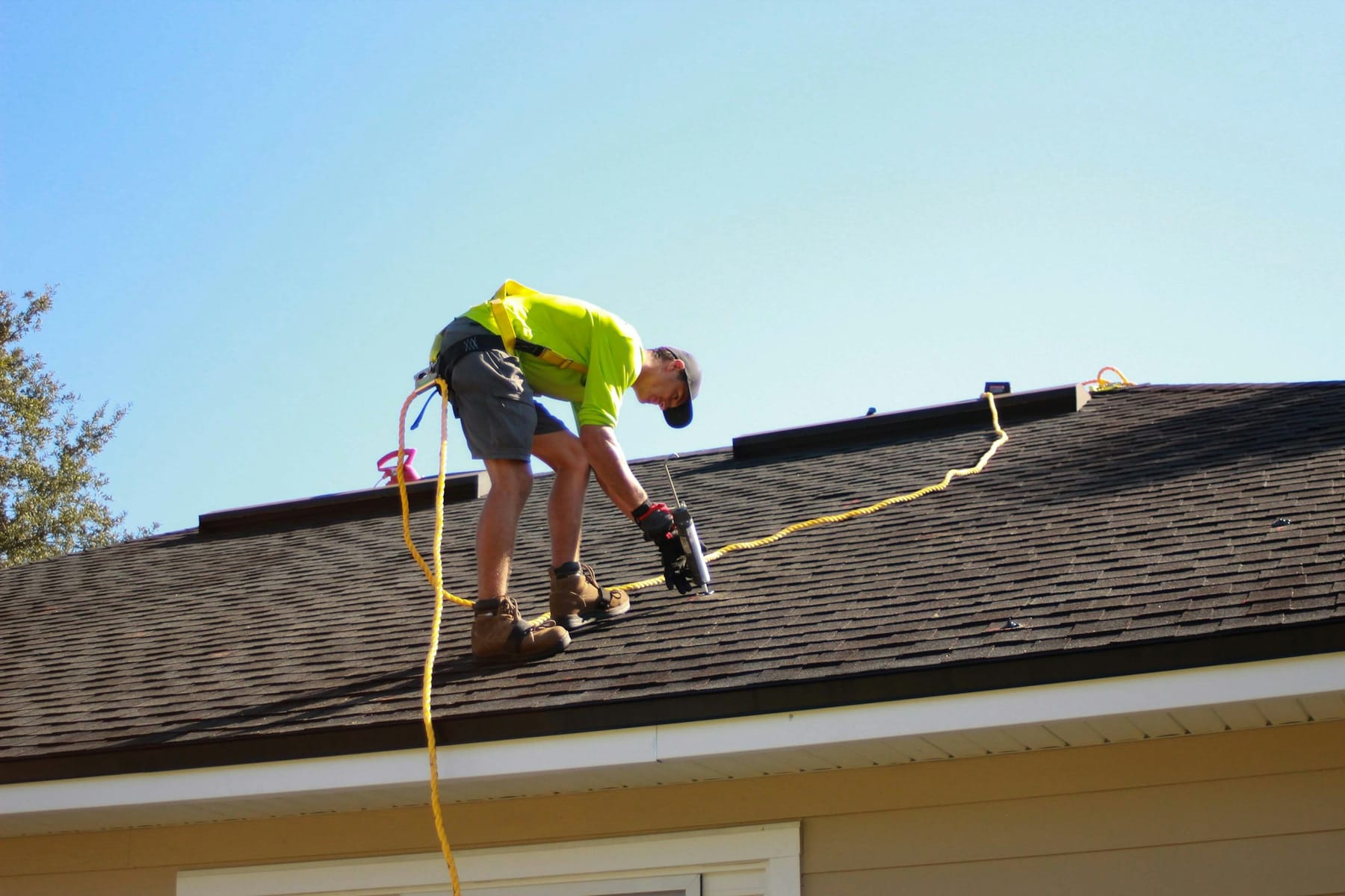 A roofer in a safety harness working on a shingle roof with a power drill